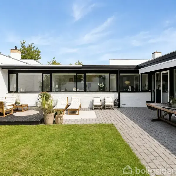 En moderne, hvid murstensbygning står roligt omkranset af buske og et træ foran huset. Sollys oplyser området, og himlen er blå med spredte skyer.