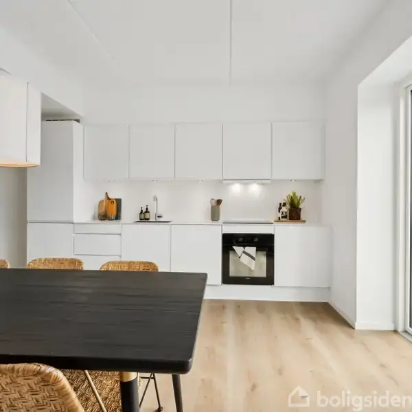 Køkkenbord med kurvestole står foran et moderne, hvidt køkken med indbygget ovn. Store vinduer giver lysindfald fra højre, og lyse trægulve dominerer rummet.