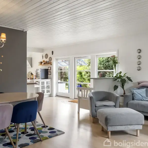 Stue med sofa, lænestol og sofabord til højre. Rundt spisebord med farverige stole til venstre. Lysekrone i loftet. Store vinduer viser grøn have udenfor.