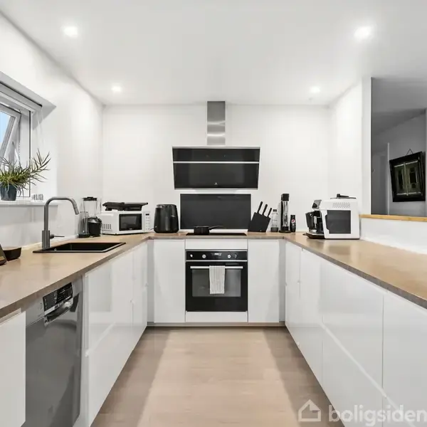 Et moderne køkken med hvide skabe og træbordplader. En indbygget ovn og komfur er centralt placeret, flankeret af forskellige køkkenapparater. Et stort vindue til venstre lader lys ind.