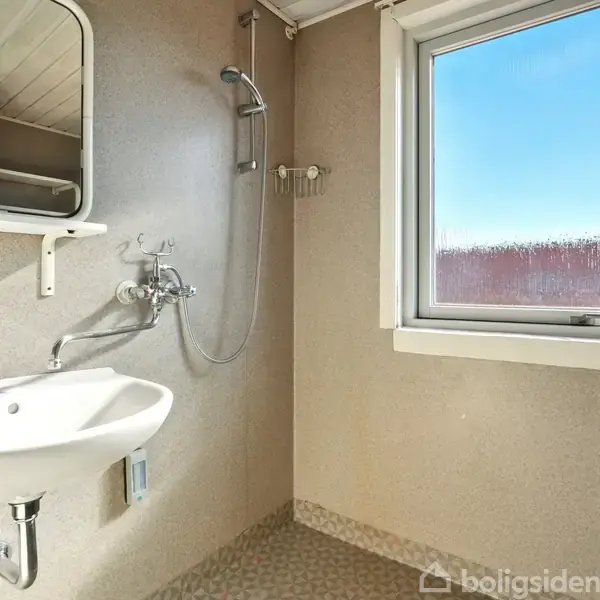 Håndvask under spejl med sæbeholder; bruser ved siden af. Vindue til højre, lys belysning. Badeværelse med simple, funktionelle faciliteter.