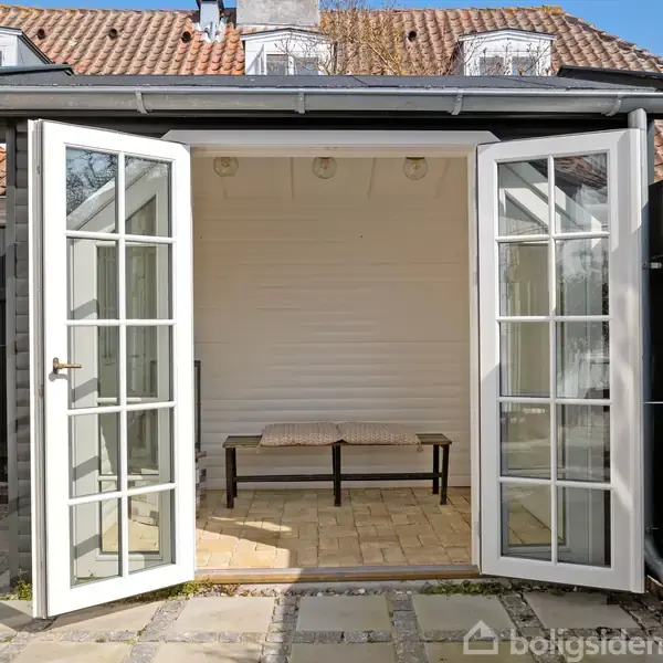 Et lille skur med åbne franske døre står i en gårdhave med flisebelægning. En bænk er placeret indvendigt, omgivet af et hegn og et hus med rødt tegltag i baggrunden.