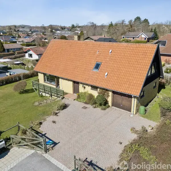 Hus med skråt, rødt tegltag og cremefarvet facade, stående på en flisebelagt indkørsel. Det er omgivet af velplejede græsplæner og beliggende i et villakvarter med flere lignende huse.