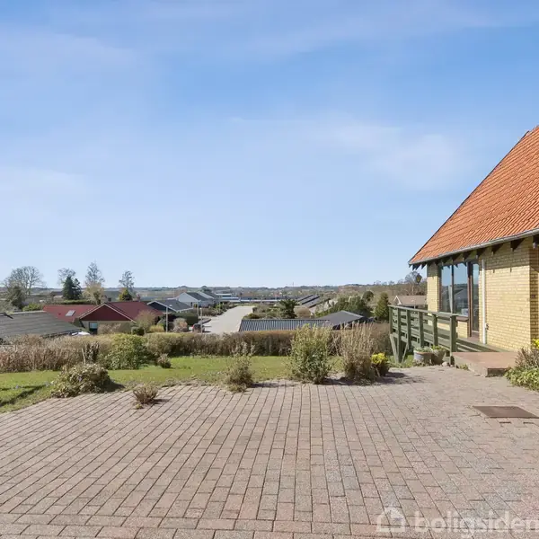 Hus med gult murværk og rødt tegltag står stille i forgrunden. Omgivet af brolagt gårdsplads, med udsigt over et villakvarter og grønne træer i baggrunden.