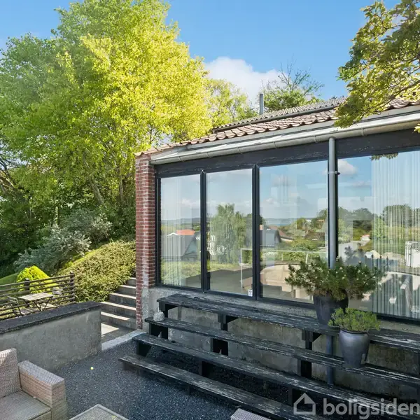 En lille tilbygning med store vinduer reflekterer lys. Den står på en terrasse med træmøbler og udsigt til en grøn have og klare himmel.