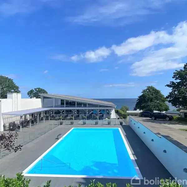 En stor, rektangulær swimmingpool står tom under en klar blå himmel, omgivet af en terrasse. I baggrunden ses en moderne bygning og træer samt havet i horisonten.