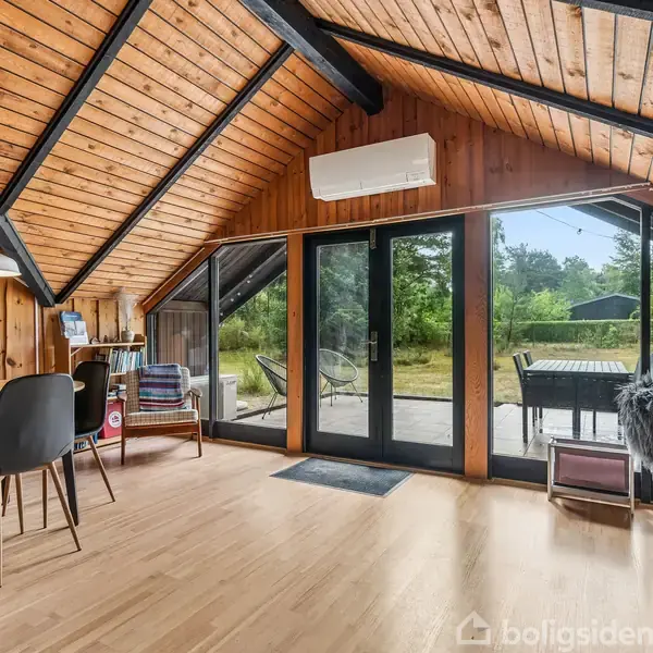 Stue med skråningetag og trævægge. Bord og stole ved venstre side, sofa ved bagvæg. Glasdøre leder ud til terrasse med bord og græsplæne i baggrunden.