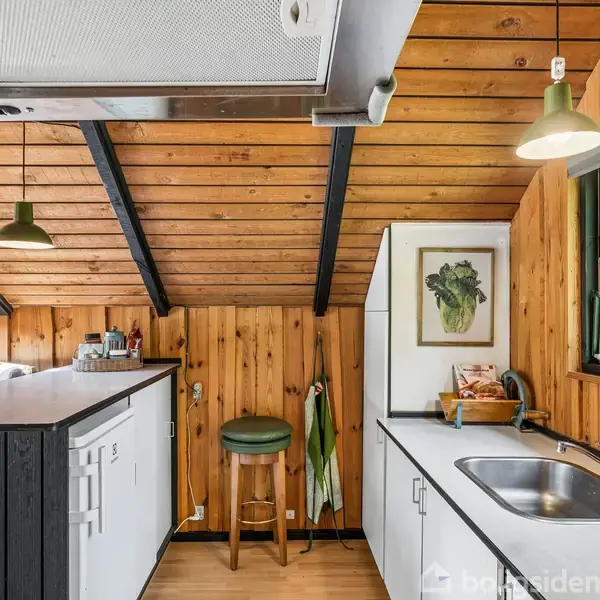 Køkken med træpanelvægge og skrå loft, indeholdende en hvid køkkenø, vask og et vindue med udsigt til grønne træer. To grønne lamper hænger fra loftet.