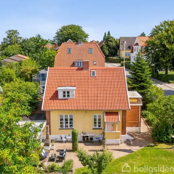 Et gult hus med rødt tegltag står i en velplejet have. Der er en terrasse med havemøbler foran huset. Til højre står en flagstang med Dannebrog.