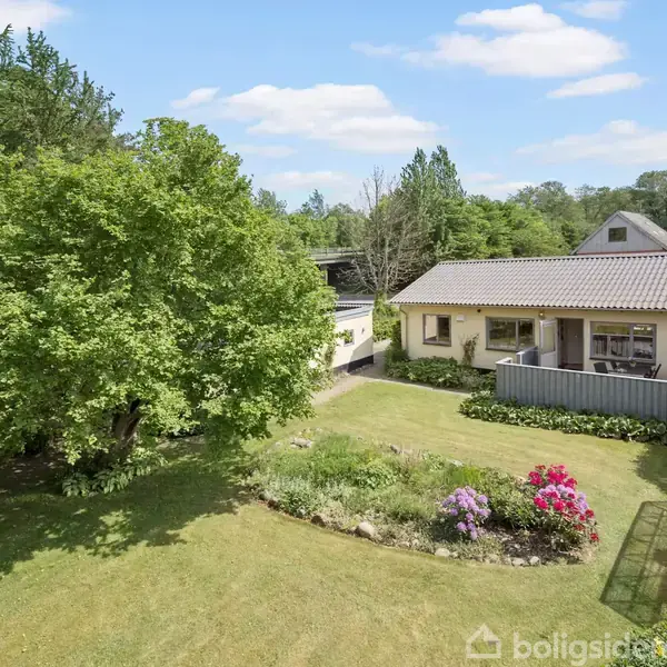 Et gult hus med terrasse står omgivet af en velholdt have med blomsterbede og et stort træ. En lille pavillon er synlig i forgrunden under en blå himmel.