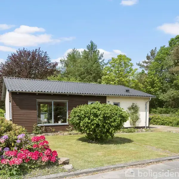 Et lille hus med fladt tag står i en have med farverige blomster og grønne buske. Rundt om huset ses en frodig, træbevokset baggrund under en blå himmel med skyer.
