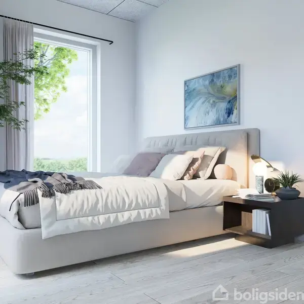 En dobbeltseng med puder og tæpper står i et lyst, moderne soveværelse. Ved siden af sengen er et natbord med en lampe og dekoration. Et stort vindue viser grønne træer udenfor.