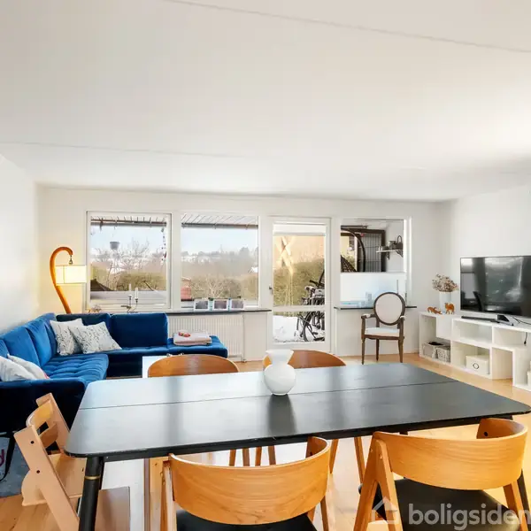Blåt sofaarrangement står i en stue med spisebord, træstole, og fladskærms-tv. Store vinduer giver udsigt til natur og en minimalistisk stil med lyse toner dominerer rummet.