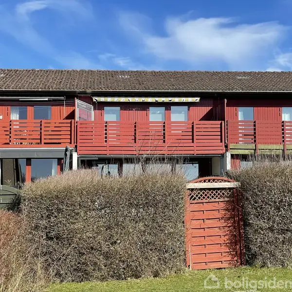Rødt rækkehus med træterrasse og flere vinduer under en blå himmel. Hækken i forgrunden er trimmet og adskiller haven fra huset.