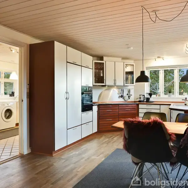 Et køkken med hvide skabe og mørke trædetaljer har en indbygget ovn og bordplads. I baggrunden ses en vaskemaskine i et tilstødende rum med flisegulv og vinduer.