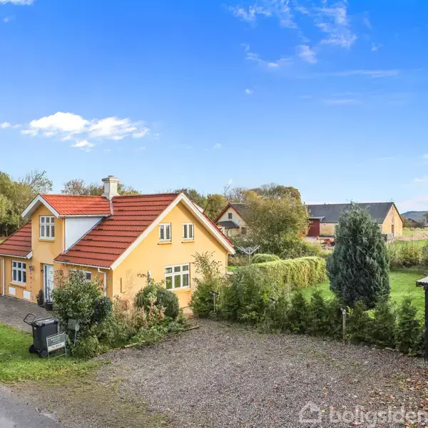 En gul to-etagers hus med rødt tegltag står omgivet af en have med buske og træer. Til højre for huset er en carport. Baggrunden viser et landligt område med flere huse.