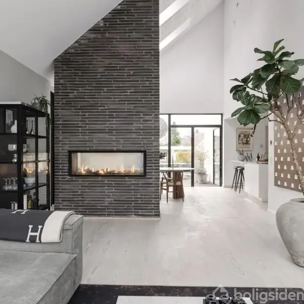 Pejs i moderne stue, omgivet af grå sofa og stort potteplante. Midtfor er en glasvæg, der fører til udendørsområde, og der er smukt dekoreret med kunst og fotos.