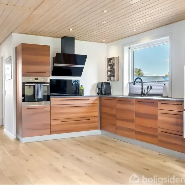 Et moderne køkken med træskabsfronter har indbyggede apparater og en sort køleskab. Store vinduer lader naturligt lys ind, og trægulve samt loft skaber en lys atmosfære.