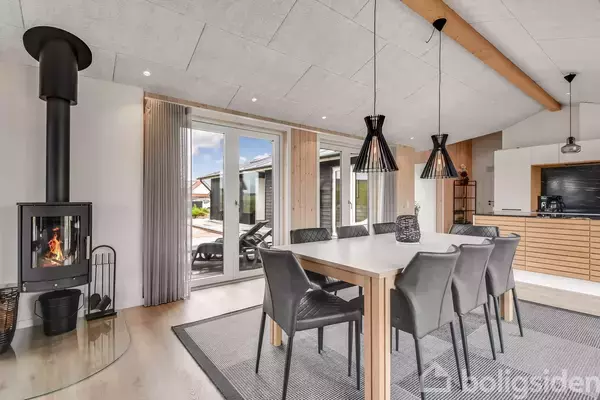 Et lyst spisestueområde med et langt bord omgivet af seks stole. Til venstre står en brændeovn med ild i. Rummet har store vinduer mod en terrasse udenfor.