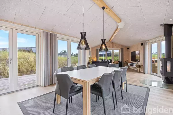 Et moderne spisebord med seks grå stole står på et tæppe. Ovenfor hænger to sorte lamper. Rummet har store vinduer og en brændeovn på højre side.