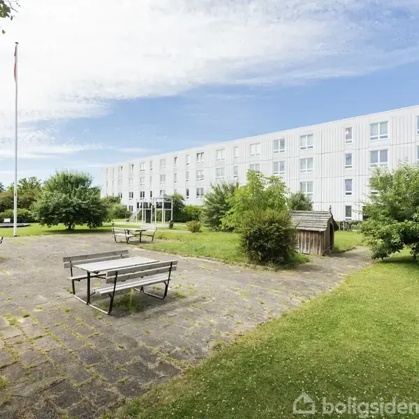 Et hvidt, moderne lejlighedskompleks står i baggrunden. Foran er der en flisebelagt gårdhave med bænke og borde, omkranset af grønne træer og en flagstang i parklignende omgivelser.