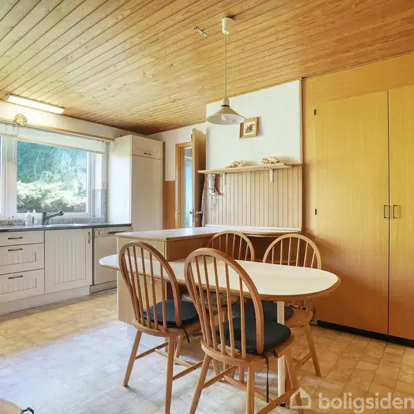 Et køkken med hvide skabe og store vinduer. Et rundt bord med fire stole står i midten, omgivet af træpaneler på væggene og et træloft.
