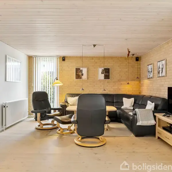 En stue med sort lædersofa og sort lænestol arrangeret omkring et sofabord. To lamper hænger over bordet. En TV står på et træbord ved siden af. Væggene er af lyse mursten.