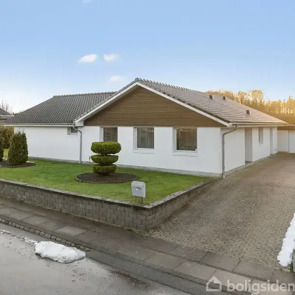 Et én-etagers hus med hvid facade og brun gavl står ved siden af en indkørsel. Foran er en trimmet busk på en græsplæne omkranset af en lav mur.