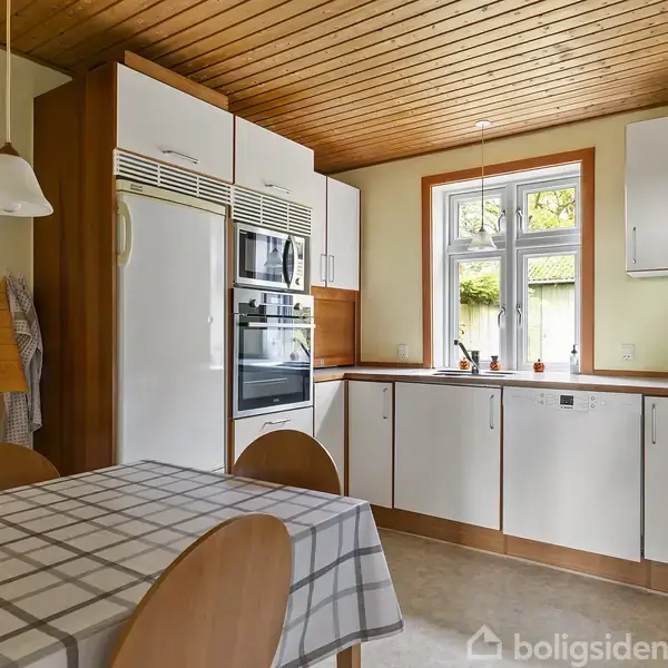 Spisebord dækket med ternet dug står i køkken med træloft. Køleskab, ovne og hvide skabe står tæt på vindue med udsigt til have. Lys hænger over bordet.