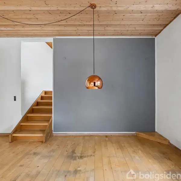 En lampe hænger fra et trælignende loft. Trappen til venstre fører til en anden etage. Væggene er hvide og grå. Gulvet er af træ. Ingen tekst.