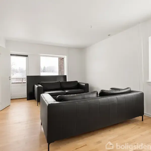 To sorte sofaer står i en stue med lyse trægulve og hvide vægge. Flere vinduer giver et lyst rum med udsigt over et udendørs område.