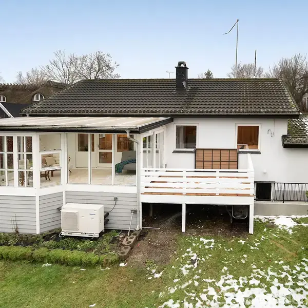 Et hvidt hus med sort tag står i en have med lidt sne på græsset. En aflukket terrasse er vedhæftet til huset, omgivet af træer og buske.