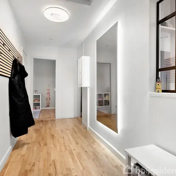 En lang gang med træparketgulv; en sort jakke hænger på knagerække. En stor spejl hænger på højre væg, og et klædeskab står for enden af gangen.