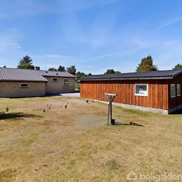 Træhus og murstenshus står i en have. Træhuset har røde vægge, mens murstenshuset er beige. Græs omkranser bygningerne, og himlen er klar og blå.