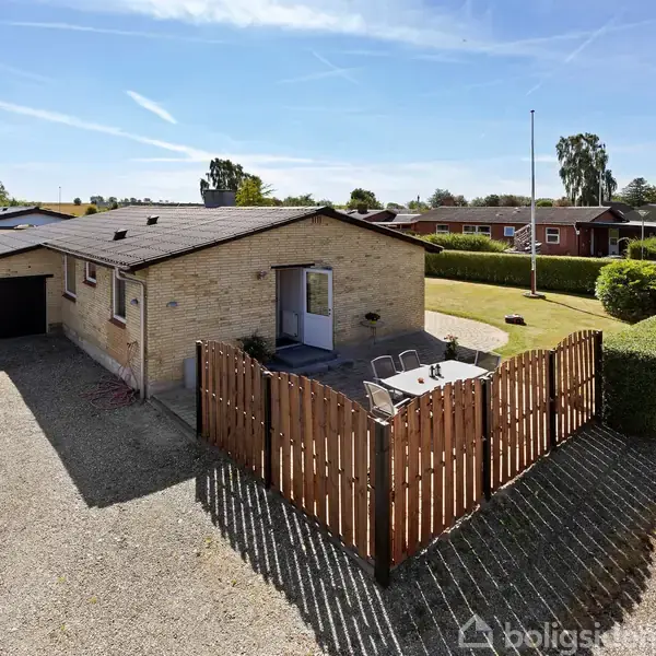 Et enkelt etplanshus i beige mursten med en indhegnet terrasse og havemøbler foran. Huset har en garage til venstre og grusbelagt indkørsel, omgivet af grønne hække og naboer.