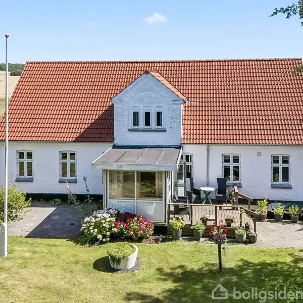 Et hus med rødt tag står stille omgivet af træer og kornmarker. Flere bygninger er i nærheden, og små køretøjer er synlige på grusvejen, der forbinder dem.