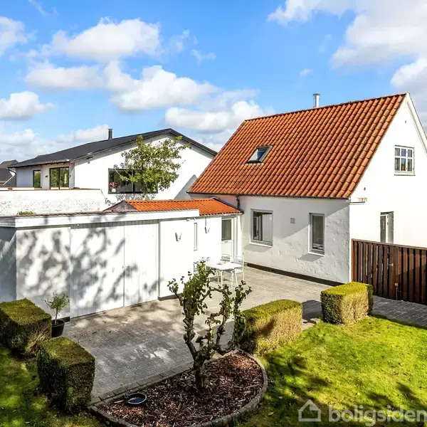 Hvidt hus med rødt tegltag står i en solrig have med grøn plæne og hække. Pår sideliggende terrasse med fliser omgives af træhegn og grønne buske. Tæt bebyggelse ses i baggrund.