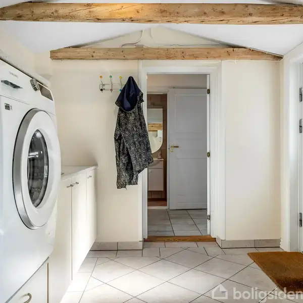 Vaskemaskine og tørretumbler står ved siden af hinanden i et lyst bryggers med synlige loftsbjælker. Frakker hænger på kroge på væggen. En dør fører til et andet rum.