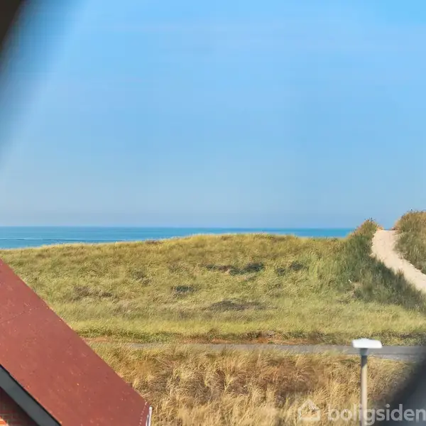 Udsigt gennem et vindue: Et tag i forgrunden, en sti fører gennem græsbeklædte klitter mod havet under en blå himmel.