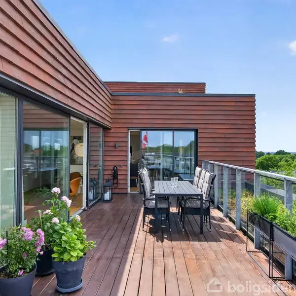 En stor tagterrasse med trægulv og glasrækværk. Møbleret med spisebord og stole omgivet af blomsterkrukker. I baggrunden ses en rød træfacade og grøn udsigt.