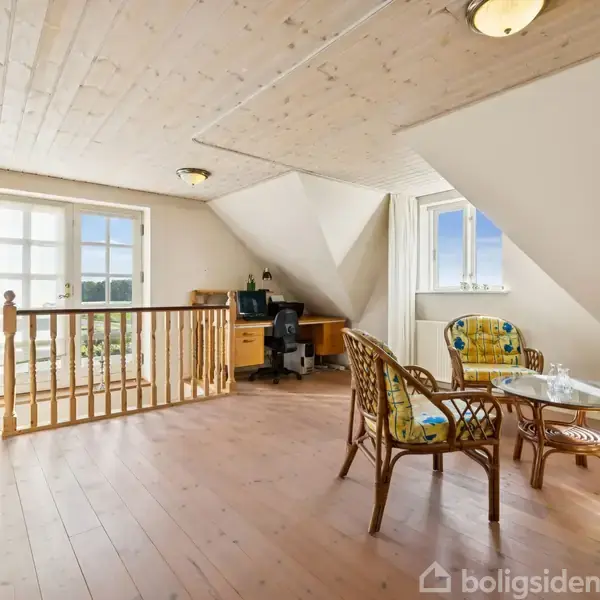 En møbleret stue på et loft med skrå vægge, indeholdende et skrivebord, computer, to polstrede stole, sofabord, og franske døre der åbner til en balkon.