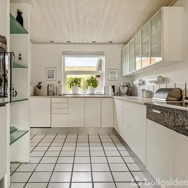 Et moderne køkken med hvide skabe og sorte granitbordplader; sollys strømmer ind fra vinduet, som har udsigt til et hus. Fliser på gulvet skaber et rent udseende.