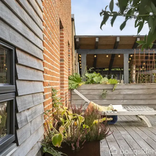 En liggestol står på en træterrasse ved siden af en murstensbygning. I baggrunden ses en pergola med grønne planter og lyskæder, omgivet af en have.