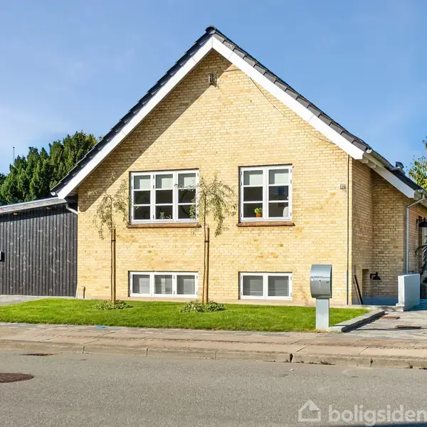 En enkelthus i gule mursten med symmetriske vinduer står på en velplejet græsplæne, omgivet af en asfaltvej og grønne træer under en klar blå himmel.