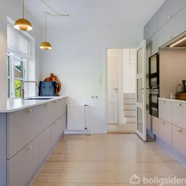 Køkken med grå skabe og indbyggede apparater. To gyldne lamper hænger over en køkkenø. Et vindue ved siden af en dør, der fører til en hvid korridor.