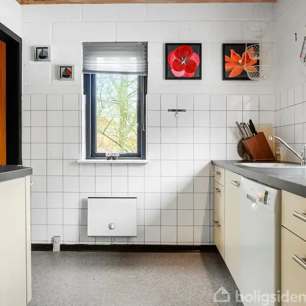 Køkken med hvide skabe og grå bordplader, stille i et rum med hvide fliser på væggene. Et vindue med persienner og farverige billeder er placeret over køkkenvasken.