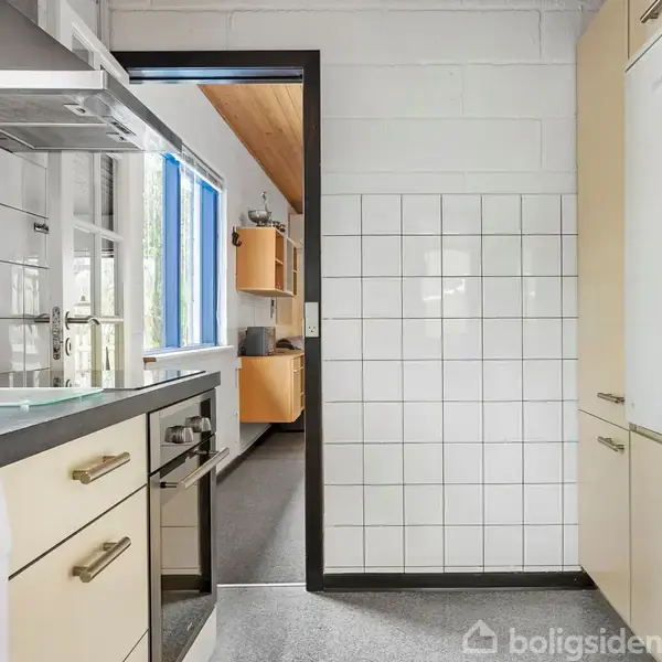 Køkken med beige skabe og sort bordplade. En opvaskemaskine fra Bosch er synlig. Gennem et åbent dørparti ses et rum med trægulv og loft. Flisebelagte vægge omkranser rummet.