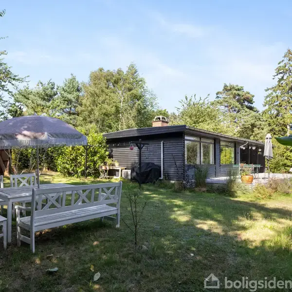 Et sommerhus omgivet af en grøn have indeholder et udendørs siddeområde med hvide træmøbler og en parasol. Skoven omkranser huset på en solrig dag.