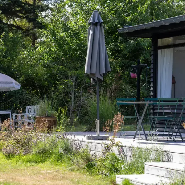 Terrassebord med parasol står foran et sort træhus. En parasol og havemøbler er placeret på græsplænen omgivet af frodig vegetation.