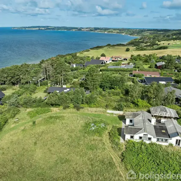 En hvid bygning med afskallet tag står i en frodig, grøn have. Den er omgivet af spredte huse og træer, med en vidstrakt udsigt over havet og skovklædte bakkedrag i baggrunden.